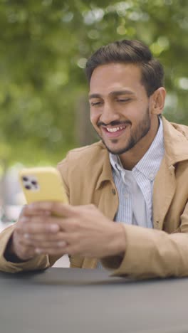 Vertikales-Video-Eines-Muslimischen-Mannes,-Der-An-Einem-Tisch-Im-Freien-Auf-Der-Stadtstraße-Sitzt-Und-Aktuelle-Textnachrichten-Auf-Dem-Mobiltelefon-Sendet